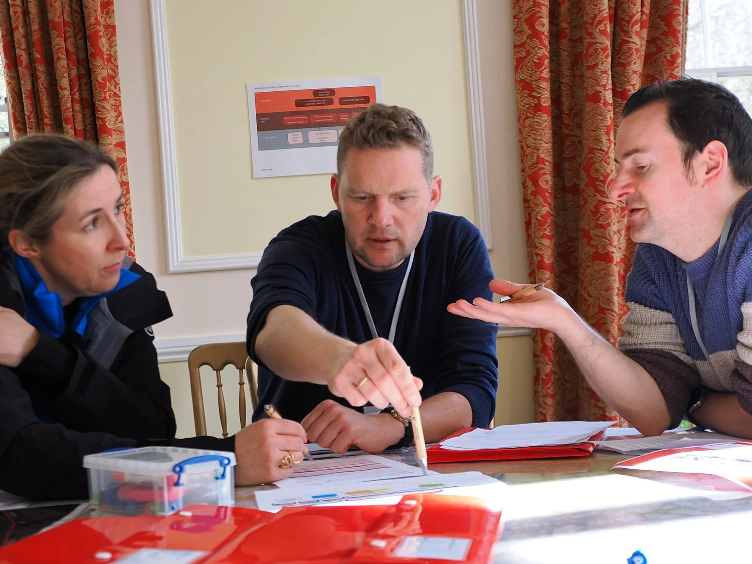 Three people sitting around a table discussing and reviewing documents, with colorful folders and papers. One person is pointing at or passing a pen over the papers.