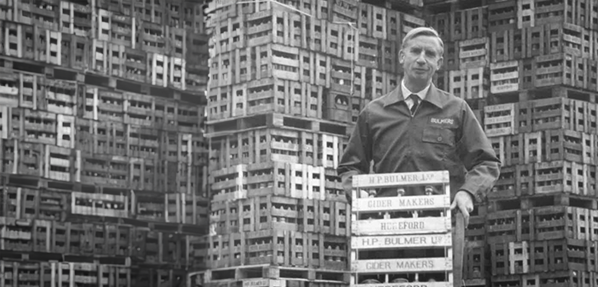 A man standing in front of stacked wooden crates, holding a tray with signs reading 'H.P. Bulmer Ltd', 'Cider Makers', and 'Hugford'.