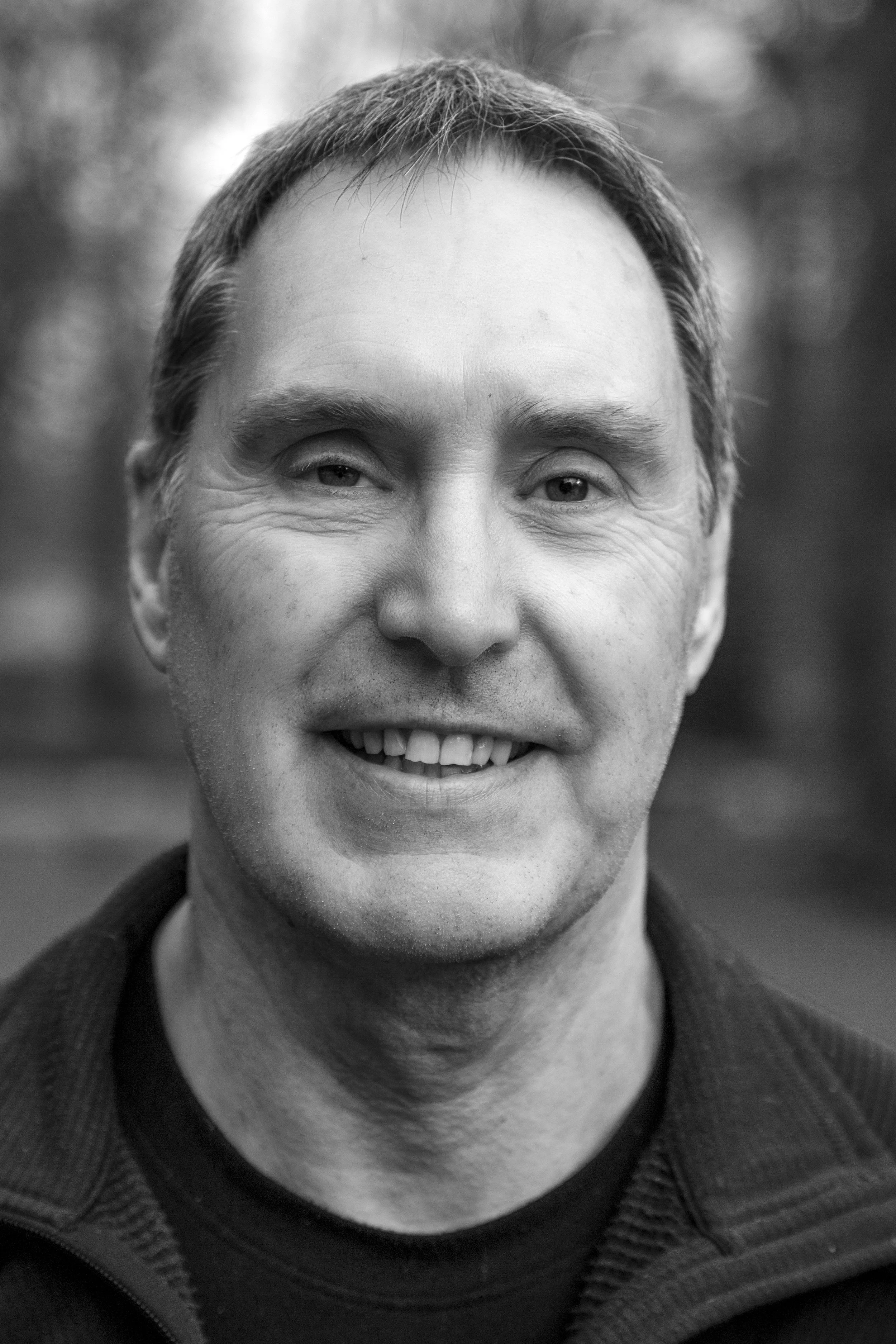 Black and white portrait of a man with short hair, smiling with teeth showing. He is wearing a zip-up jacket over a shirt, outdoors with a blurred background.