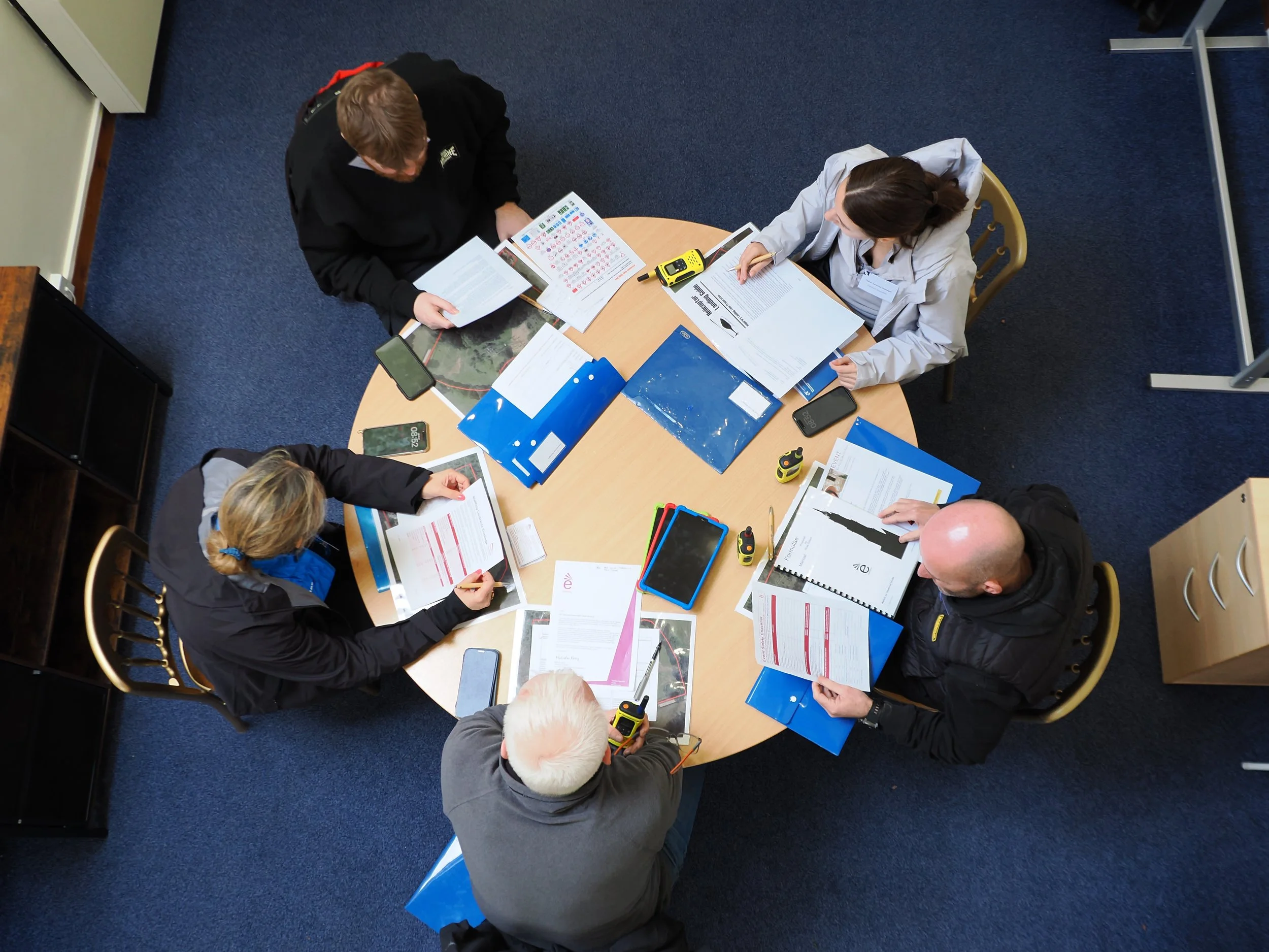Overhead view of a group of people sitting around a wooden table, engaging in discussion with various documents, pamphlets, and electronic devices on the table.