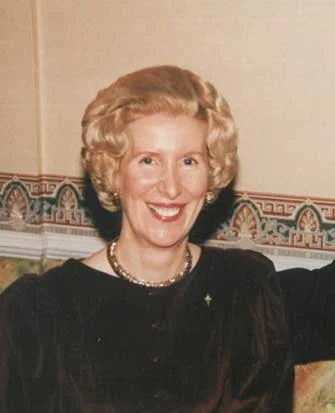 A smiling woman with curly blonde hair wearing a black dress and pearl necklace, standing indoors near a decorated wall.