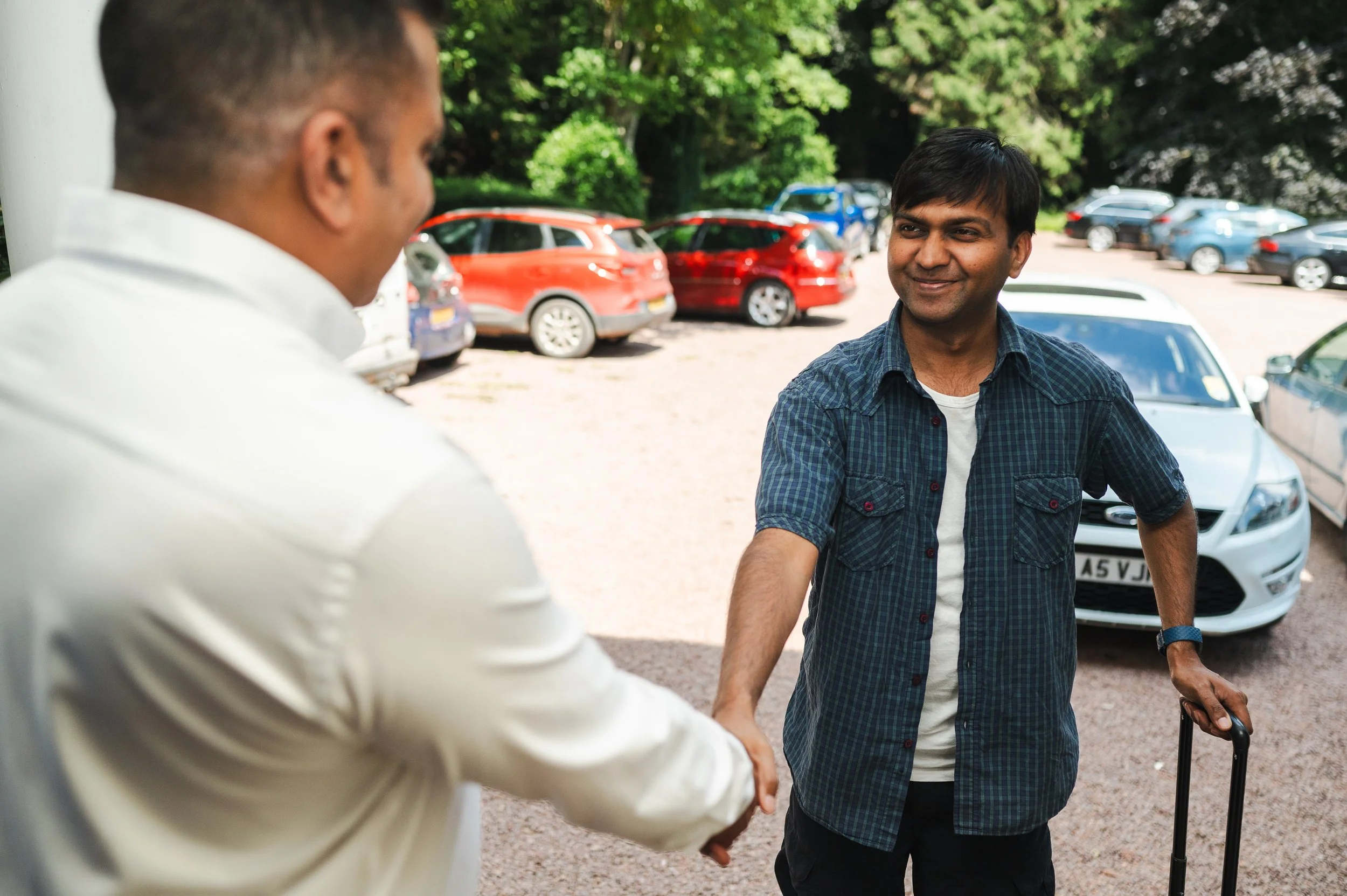 Two men are shaking hands outside in a parking lot surrounded by trees and parked cars, one man is holding a cane and smiling.