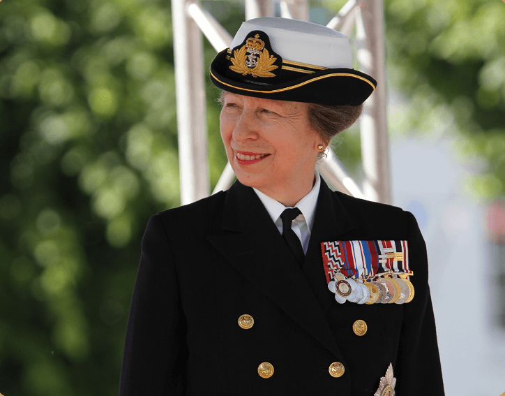 A woman in a military uniform with medals and a white hat with navy insignia, smiling outdoors.