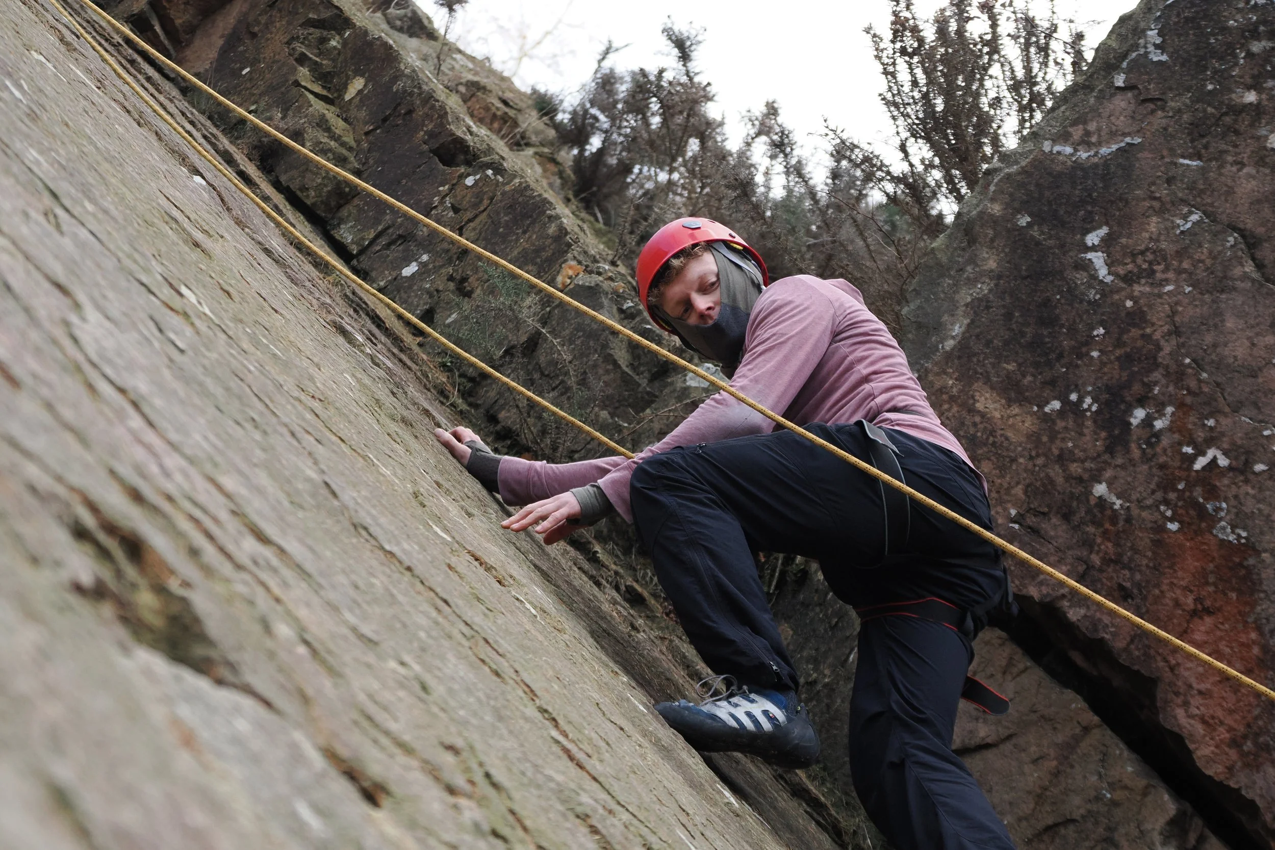 Person wearing a red helmet and blue athletic shoes rock climbing on a vertical rock face outdoors, with trees in the background.