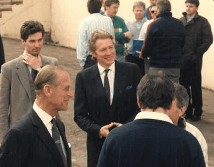 Group of men in formal and casual attire socializing outdoors, some engaged in conversation, in a setting resembling a film or TV show set.