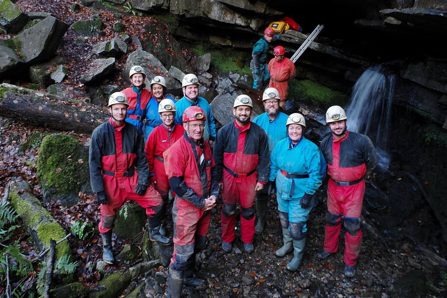 Group of people in caving gear with helmets and headlamps, standing in a rocky, cave-like area with a small waterfall in the background.