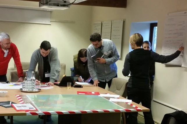 Group of professionals in a conference room engaged in a discussion, with charts on the wall and a large map on the table.