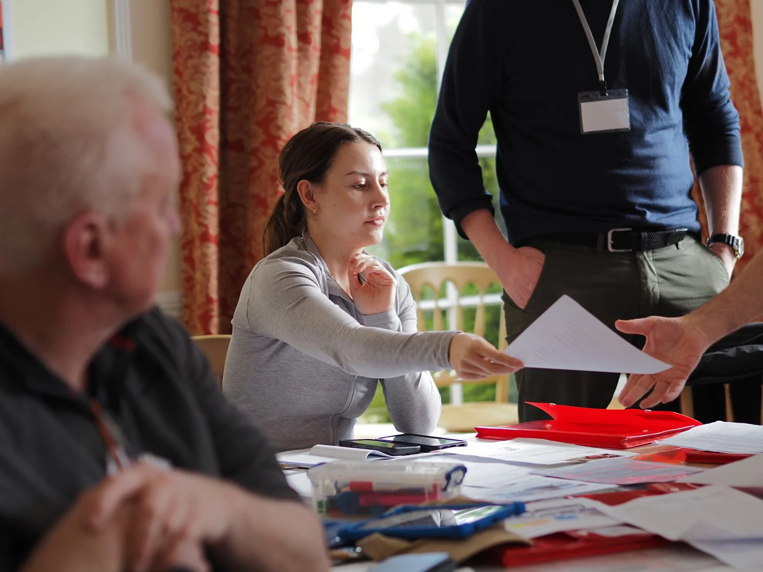 People in a meeting or a discussion, with documents, folders, and stationery on the table. A woman is passing a paper to a man, others are involved in the discussion.