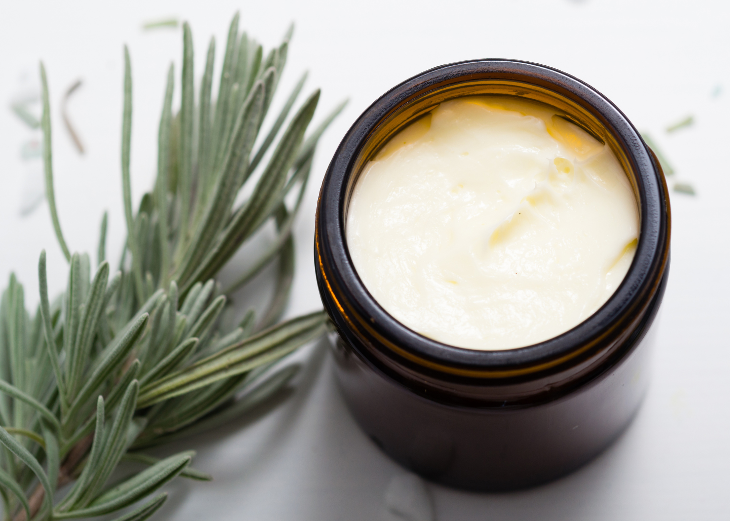 An amber glass jar filled with white balm or cream, placed on a white surface, with a sprig of fresh rosemary next to it.