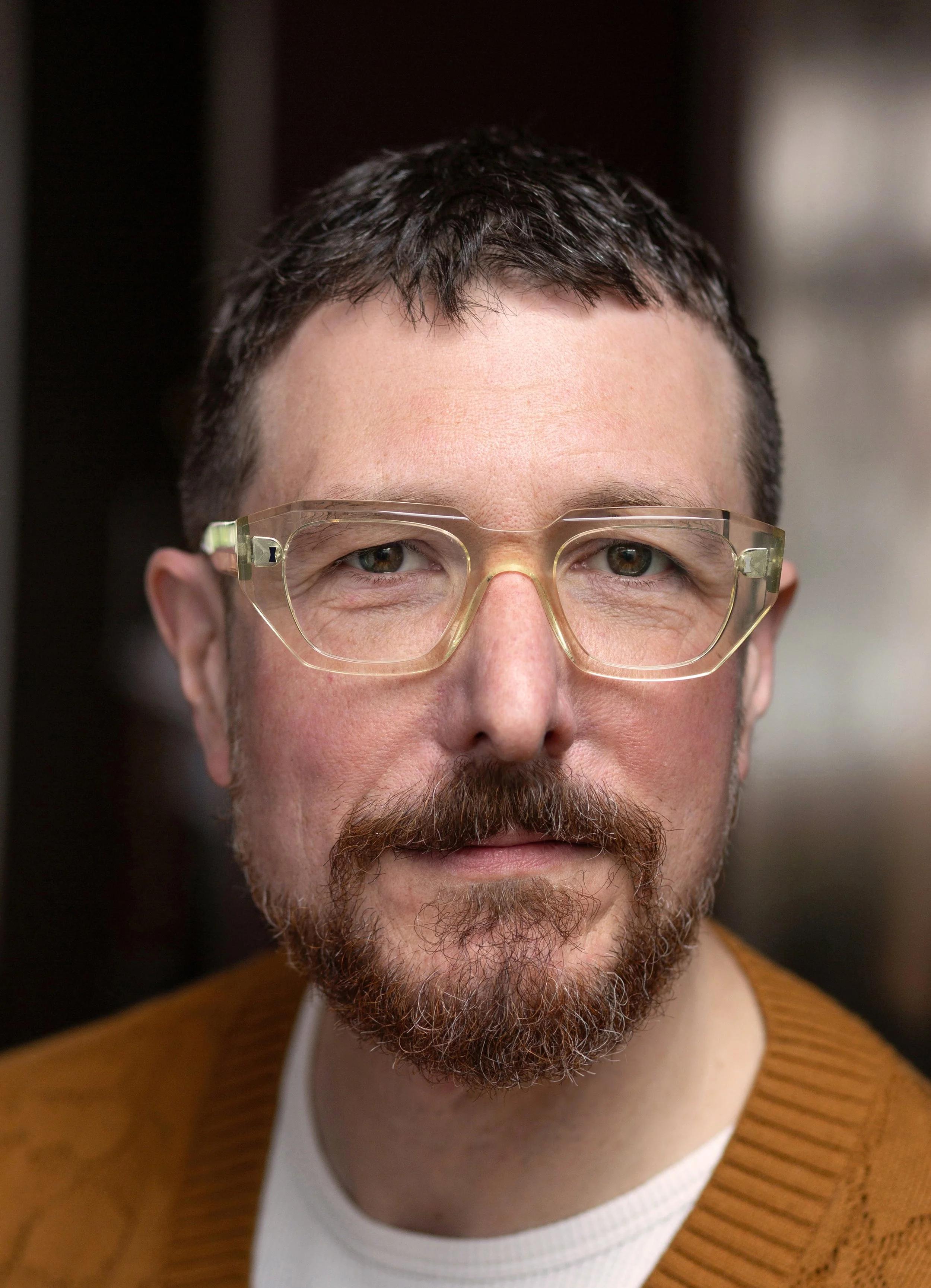 Close-up of a man with short brown hair, a beard, and glasses, wearing a brown sweater and white shirt, looking directly at the camera.