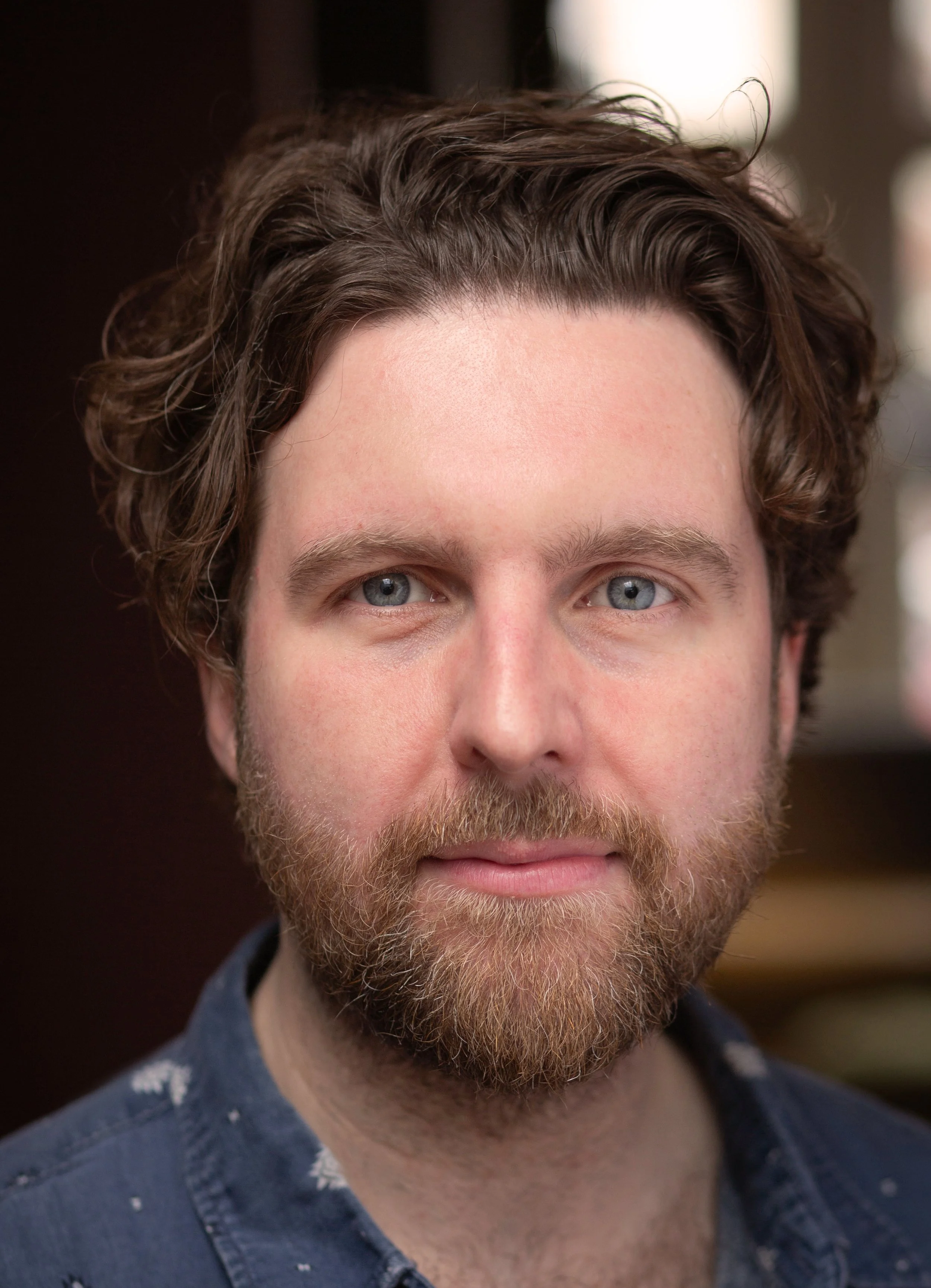 A close-up of a man's face showing blue eyes, light skin, a beard, and wavy brown hair, wearing a denim shirt.