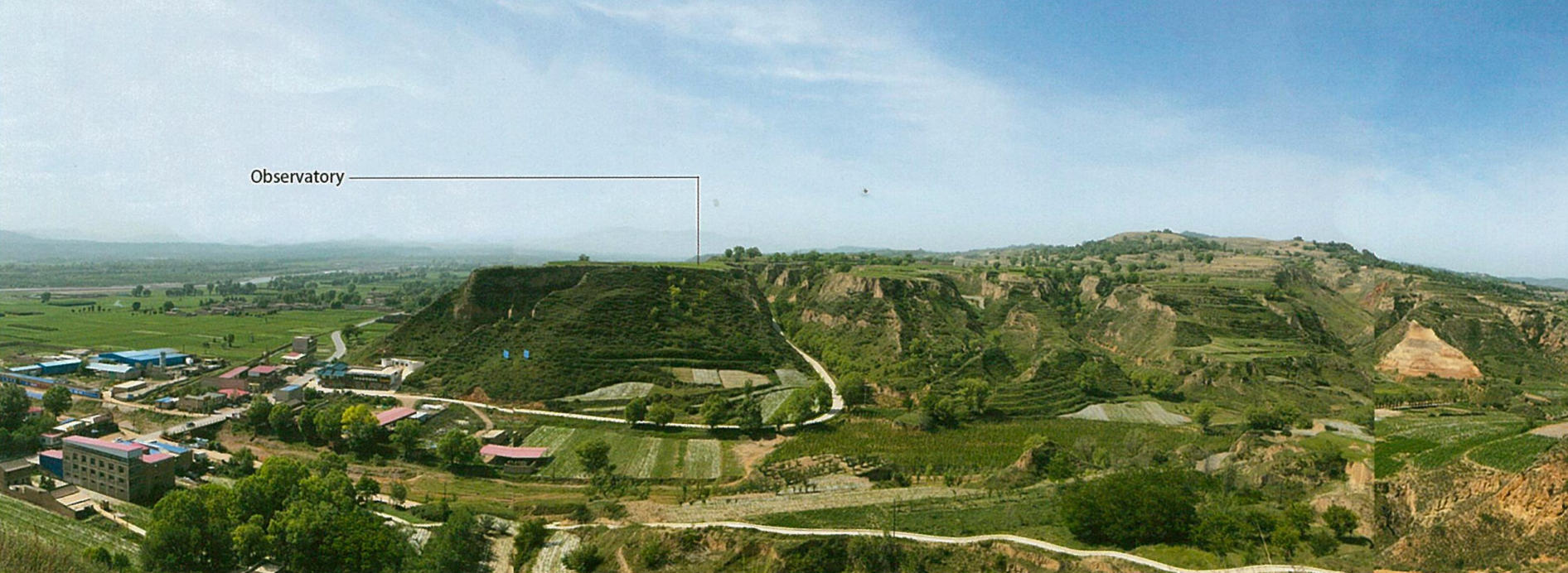 Panoramic view of a landscape with terraced hills, greenery, and a small observatory in the distance, under a partly cloudy blue sky.