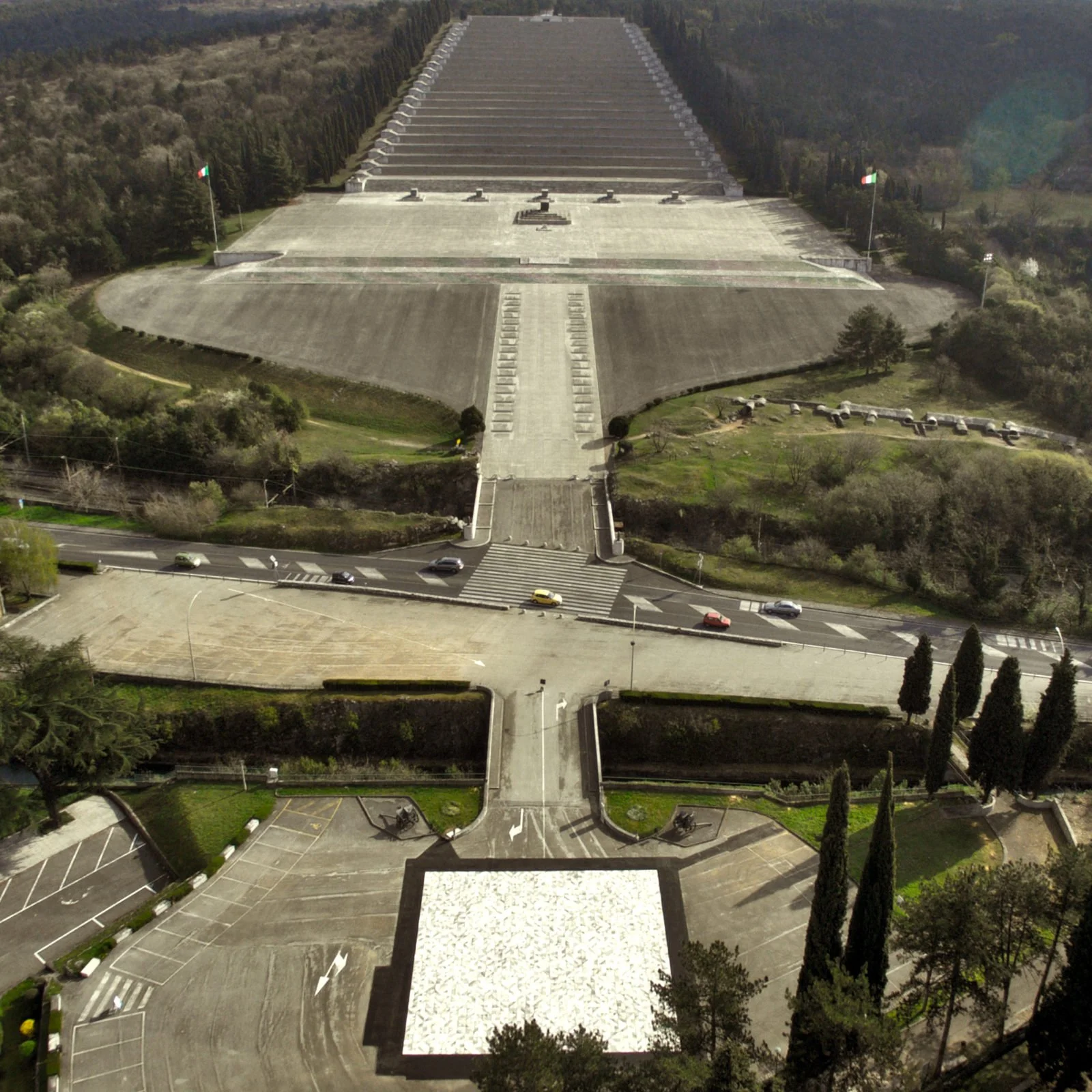 Aerial view of a large monument with tiered, wide staircase and Italian flags on either side, surrounded by greenery and roads.
