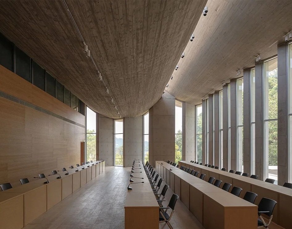 A modern lecture hall or conference room with wooden and concrete interior. It has tall, narrow windows on the right side and long tables with black chairs arranged on both sides.