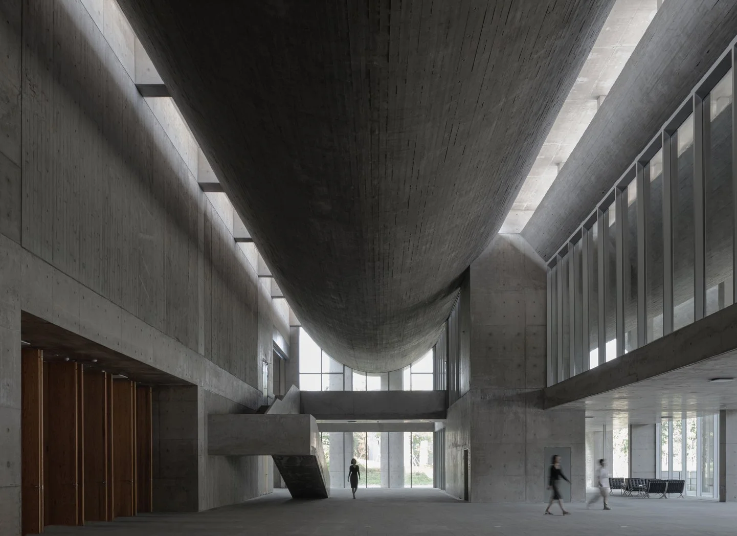 Modern interior space with concrete walls, a large cylindrical structure overhead, and large windows letting in natural light. People are walking through the space.