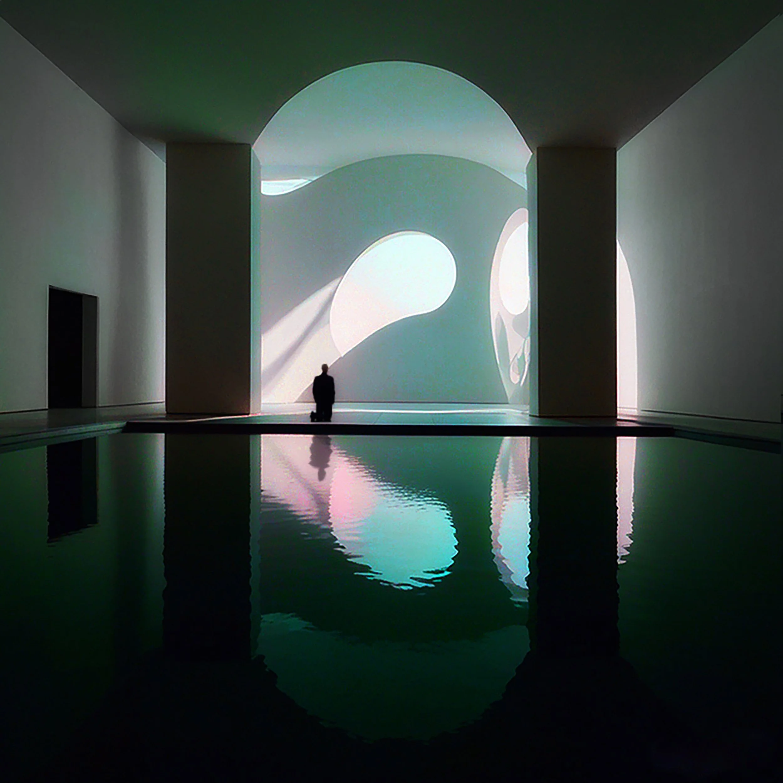 A person standing at the edge of a dark pool with illuminated abstract shapes projected on a white wall behind them.