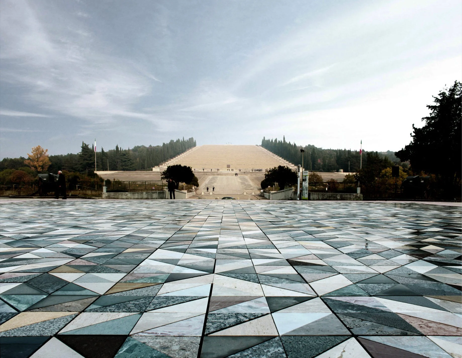 View of a large monument with wide stairs leading up, surrounded by trees and flags, possibly part of a memorial or government site, with a tiled patterned foreground. The sky is partly cloudy.