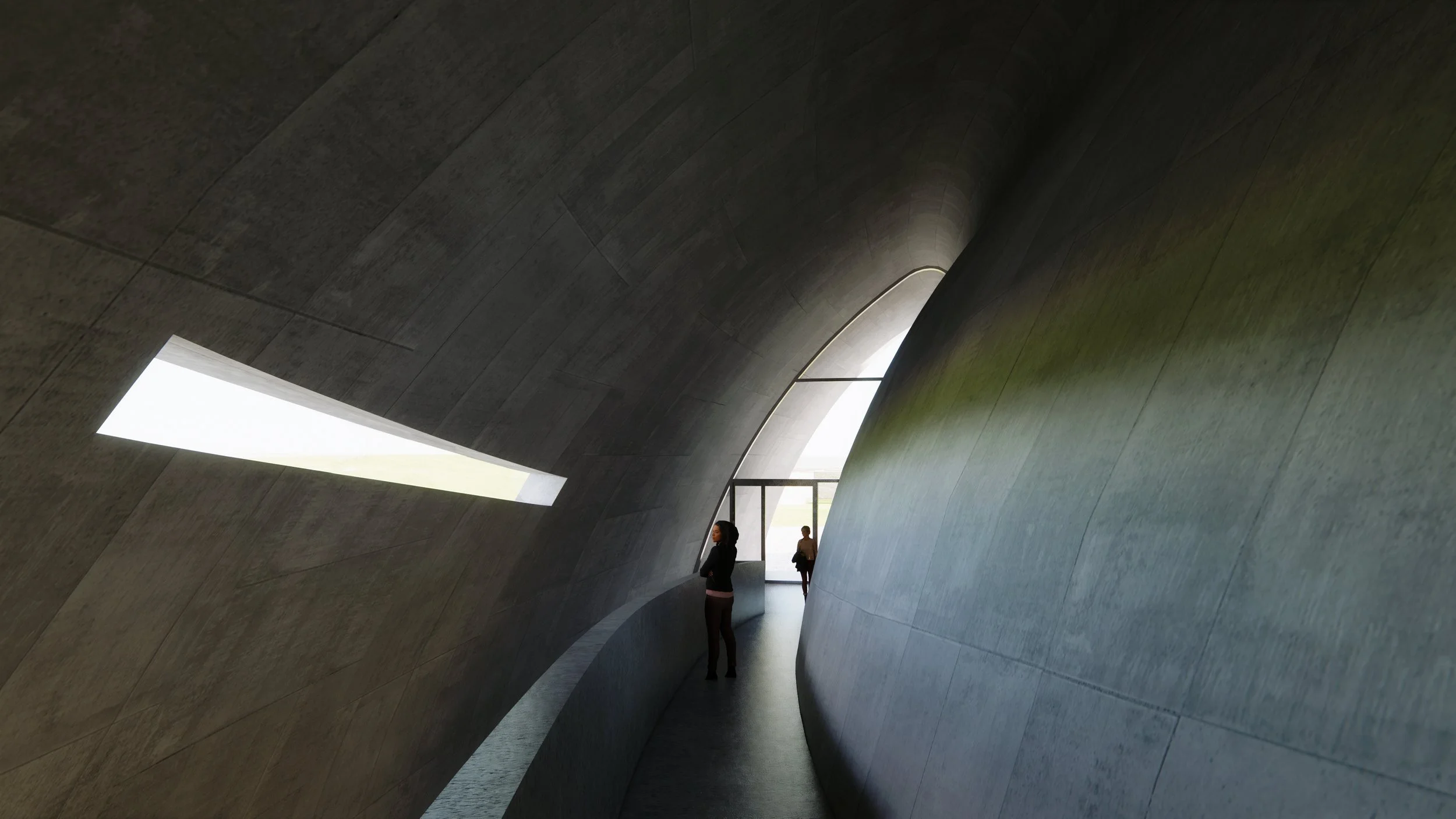 Interior of a modern building with curved, slate-gray walls and large openings letting in natural light. Two people are standing inside near an opening, one woman in black standing with her arms crossed and a man in the distance near the entrance.