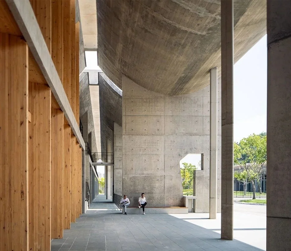 Two people sitting on a bench in a modern architectural structure with concrete walls, wooden panels, and an open outdoor area with trees.