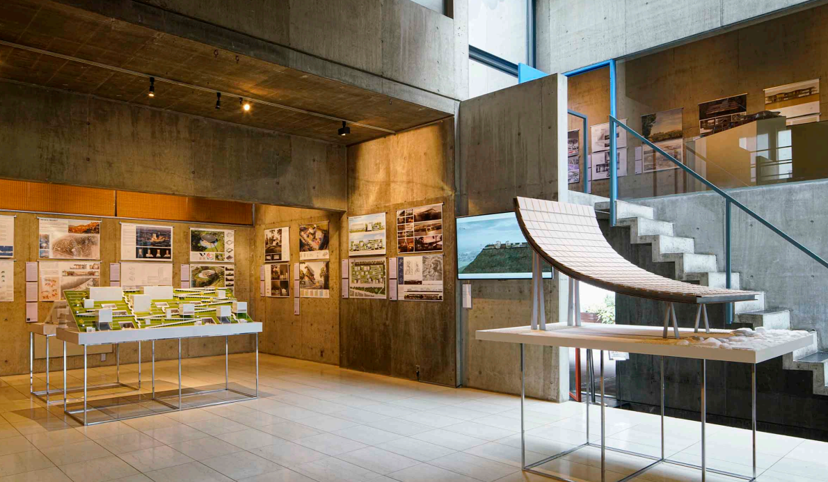 Interior of an architecture or design exhibition with models, blueprints, and photographs displayed on concrete walls and stands.