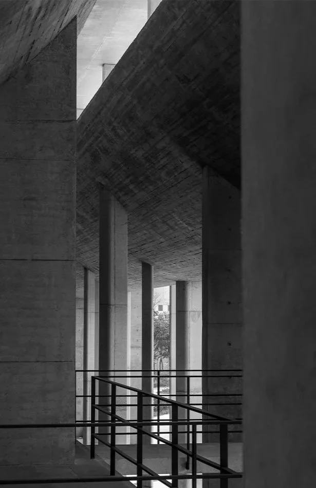 Black and white photo of a modern concrete building with tall columns and diagonal beams, featuring a railing in foreground.