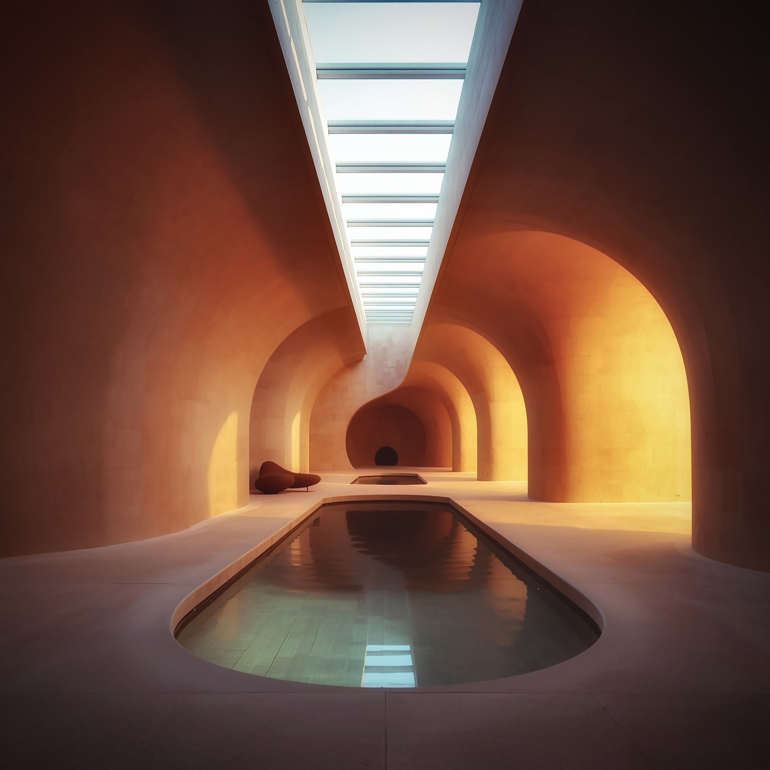 Interior of a building with a skylight at the top, curved walls painted in red, and a reflective water feature at the bottom. There are two chairs on the left side and a small table on the right side.