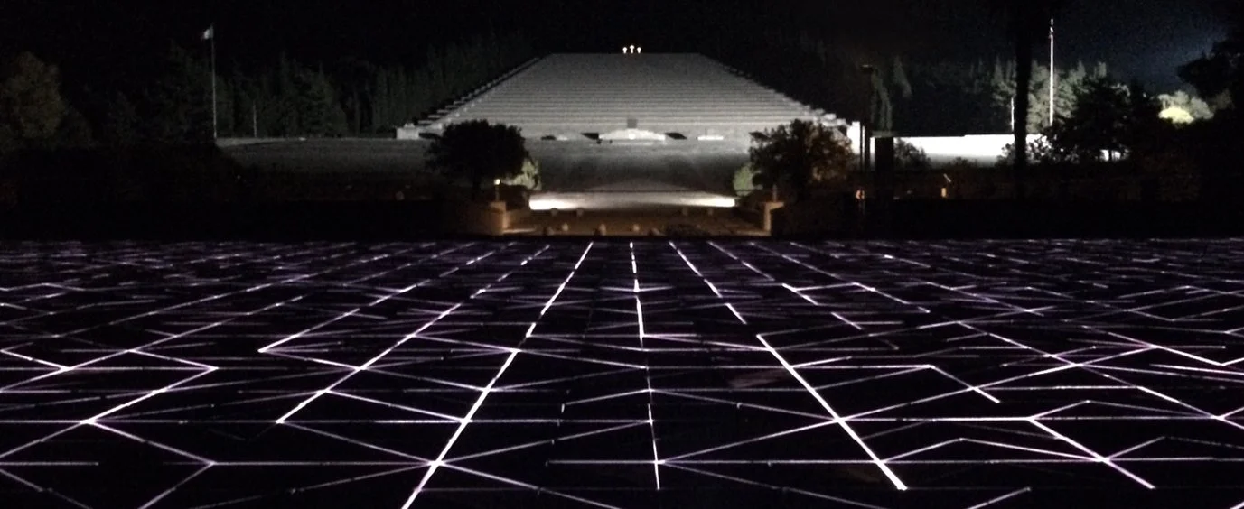 Nighttime view of a grid-like structure with glowing lines, leading to a large illuminated building in the distance.