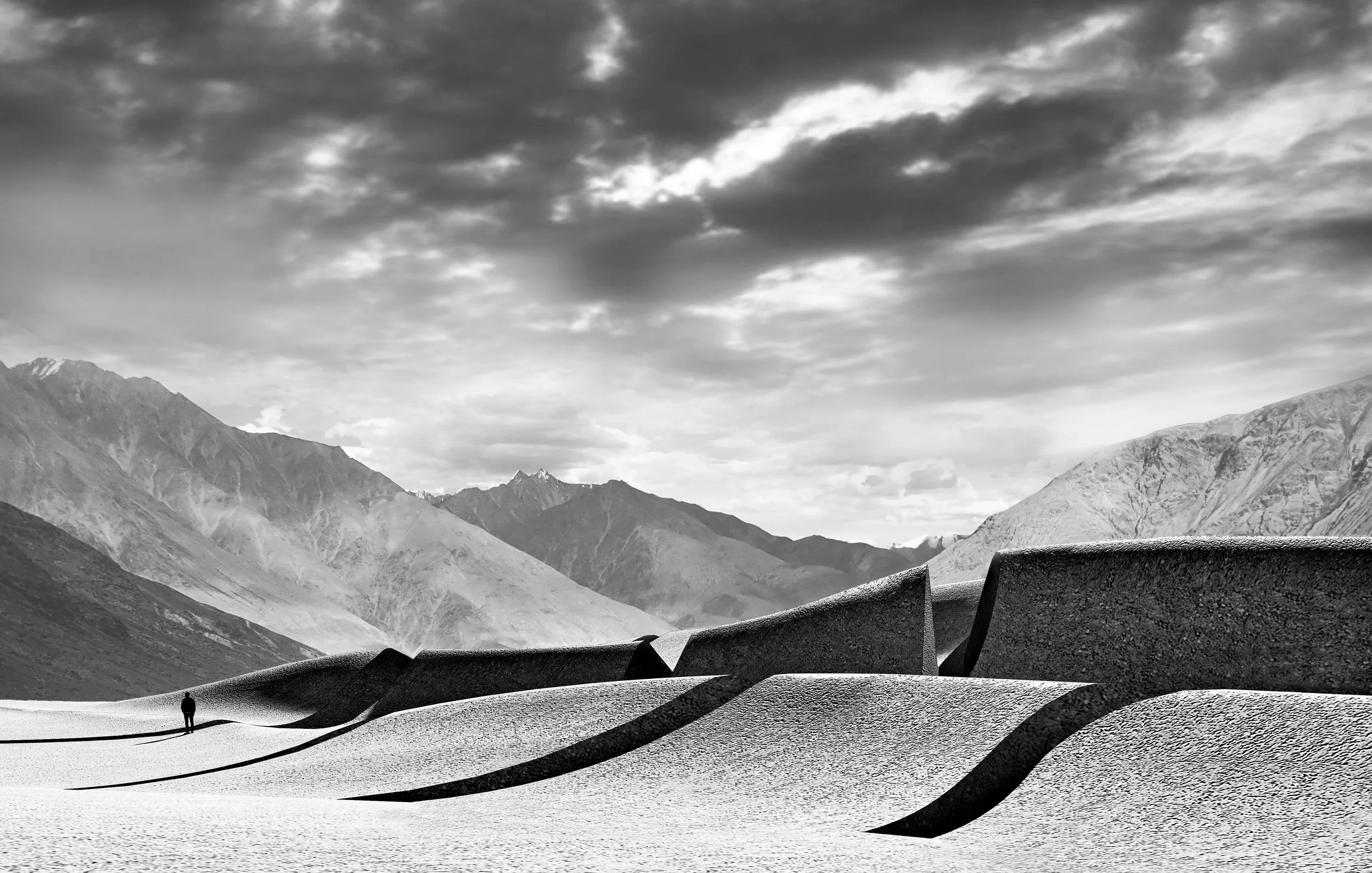 A person standing alone in a vast, snowy landscape with large mountains in the background and a cloudy sky overhead, all in black and white.