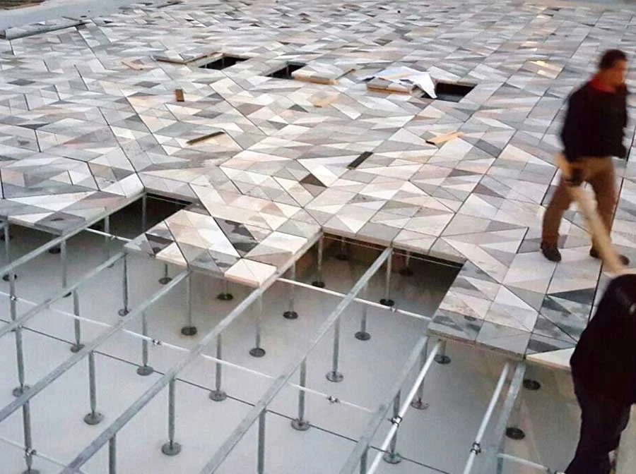 Floor with geometric tiles undergoing repair or renovation, with some tiles removed exposing the underlying structure, and a person walking past.