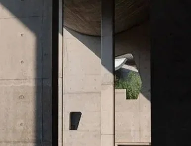A modern architectural structure with concrete walls, vertical metal beams, and an overhang casting shadow, with a glimpse of greenery in the background.