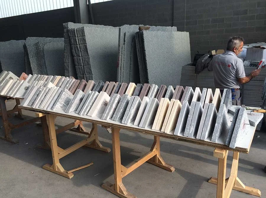 An outdoor display of various stone samples, including marble and granite, arranged on wooden tables for selection, with a man examining one of the stone pieces in the background.