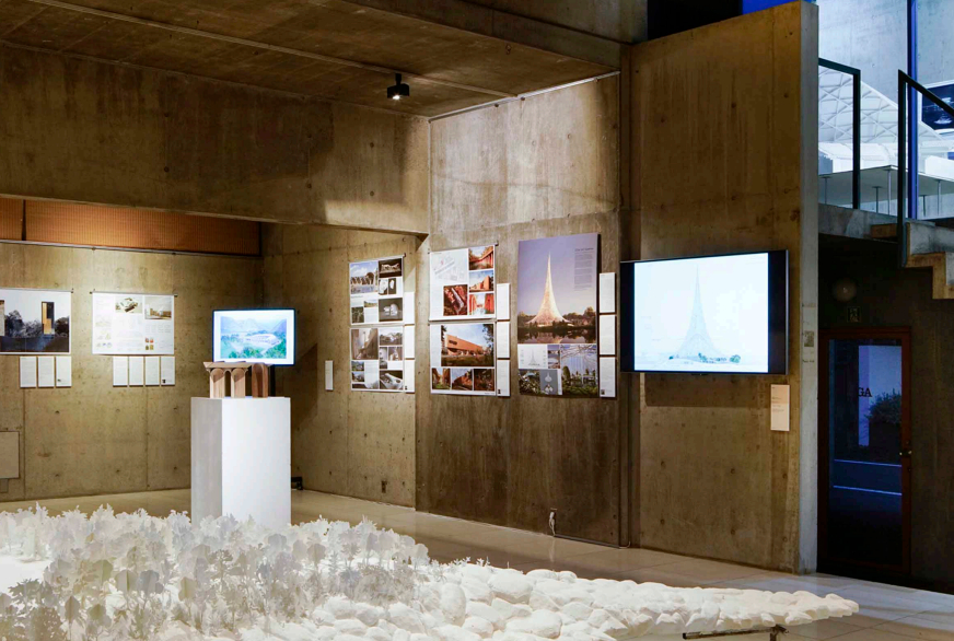 An art exhibit showcasing photographs and art pieces about the Eiffel Tower, with digital screens displaying related images, in a gallery with concrete walls and a textured white floor.