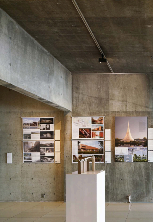 Interior of an art or architecture exhibition with concrete walls, displaying large posters of architectural projects and a sculpture on a pedestal.