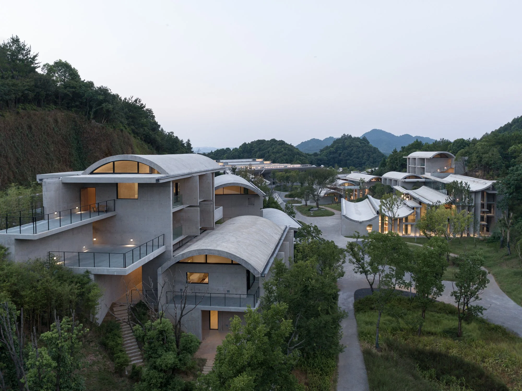 Modern multi-level buildings with curved roofs surrounded by green trees and hills in the background, during dusk.