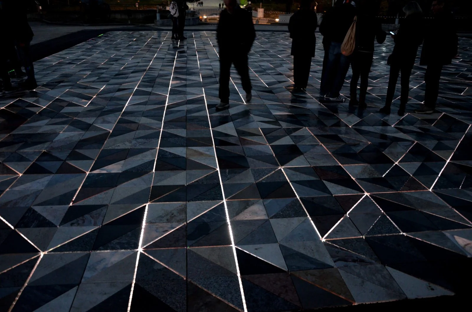 Dark tiled floor with illuminated lines forming geometric patterns, with people standing and walking in the background during evening or night.