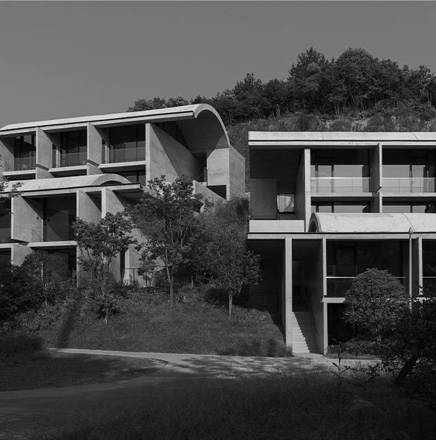 Modern multi-level concrete building with curved roof sections, large windows, and staircases, situated on a hillside with trees and greenery.
