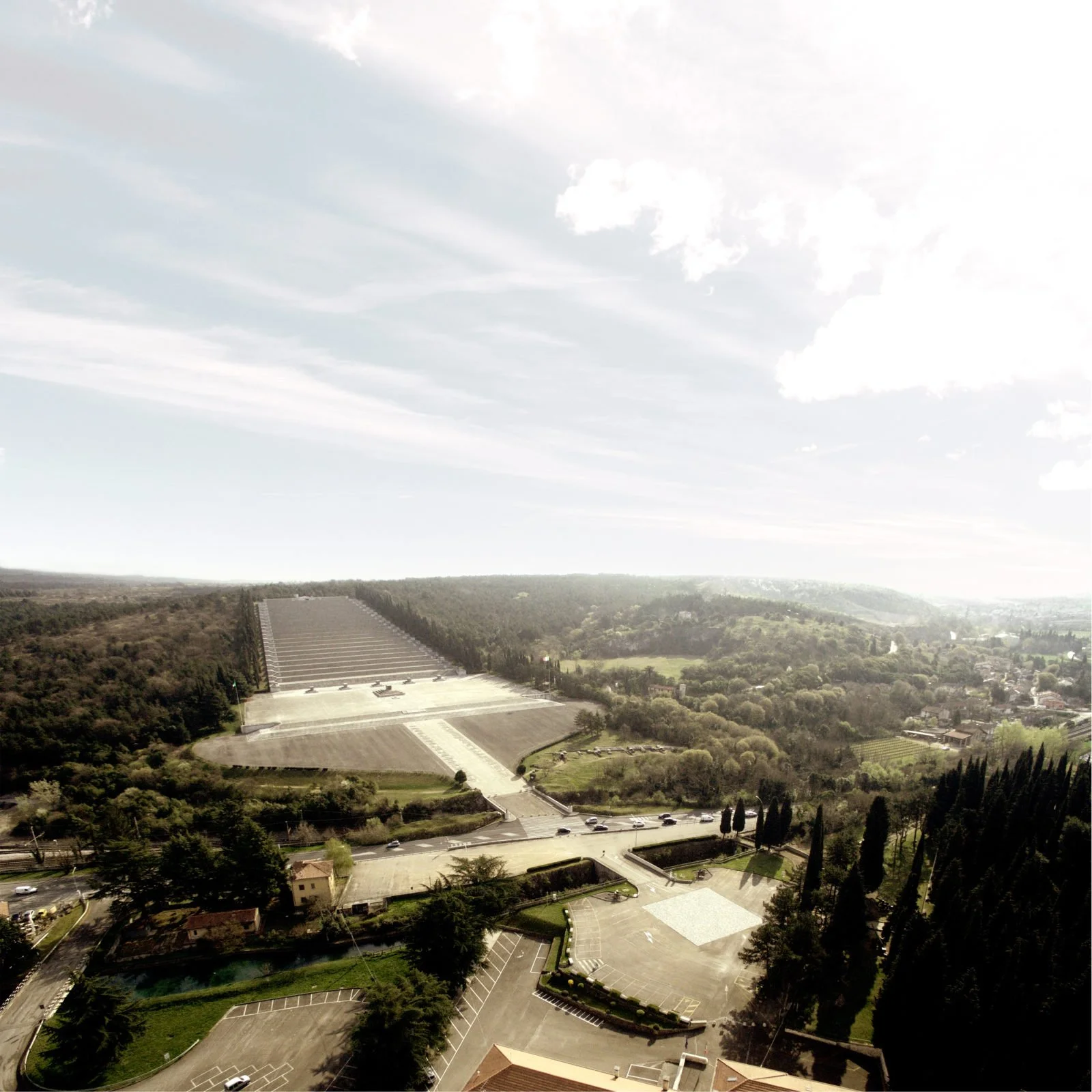 A landscape view of a countryside area with a large stairway leading up a hill, surrounded by a parking lot, trees, and fields, under a cloudy sky.