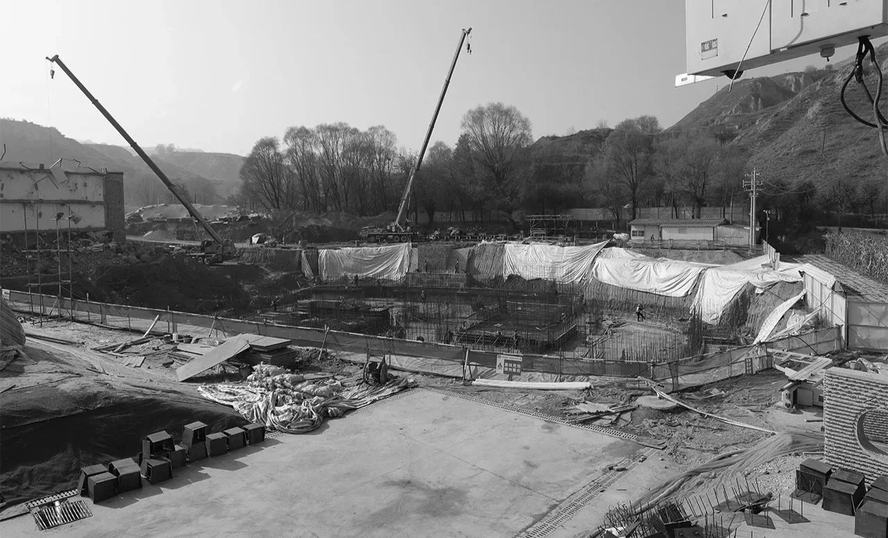 Construction site with cranes and partially built structures, surrounded by trees and hills in the background, with construction materials and equipment scattered around.