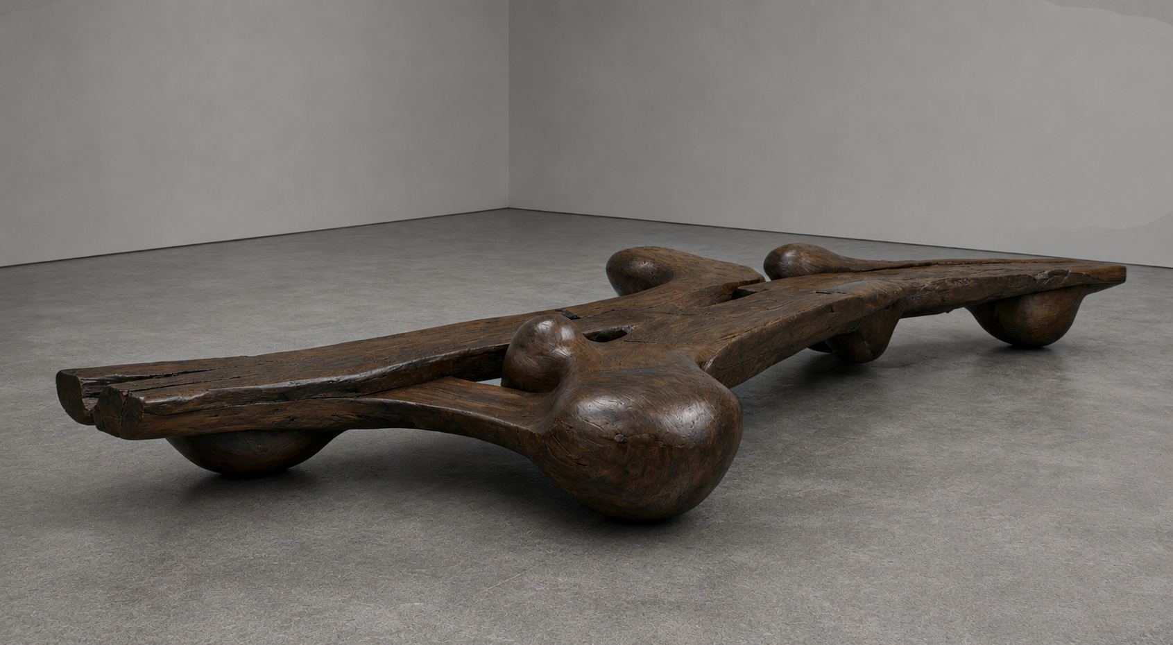 A large, irregularly shaped wooden sculpture in a gallery space with plain gray walls and floor.