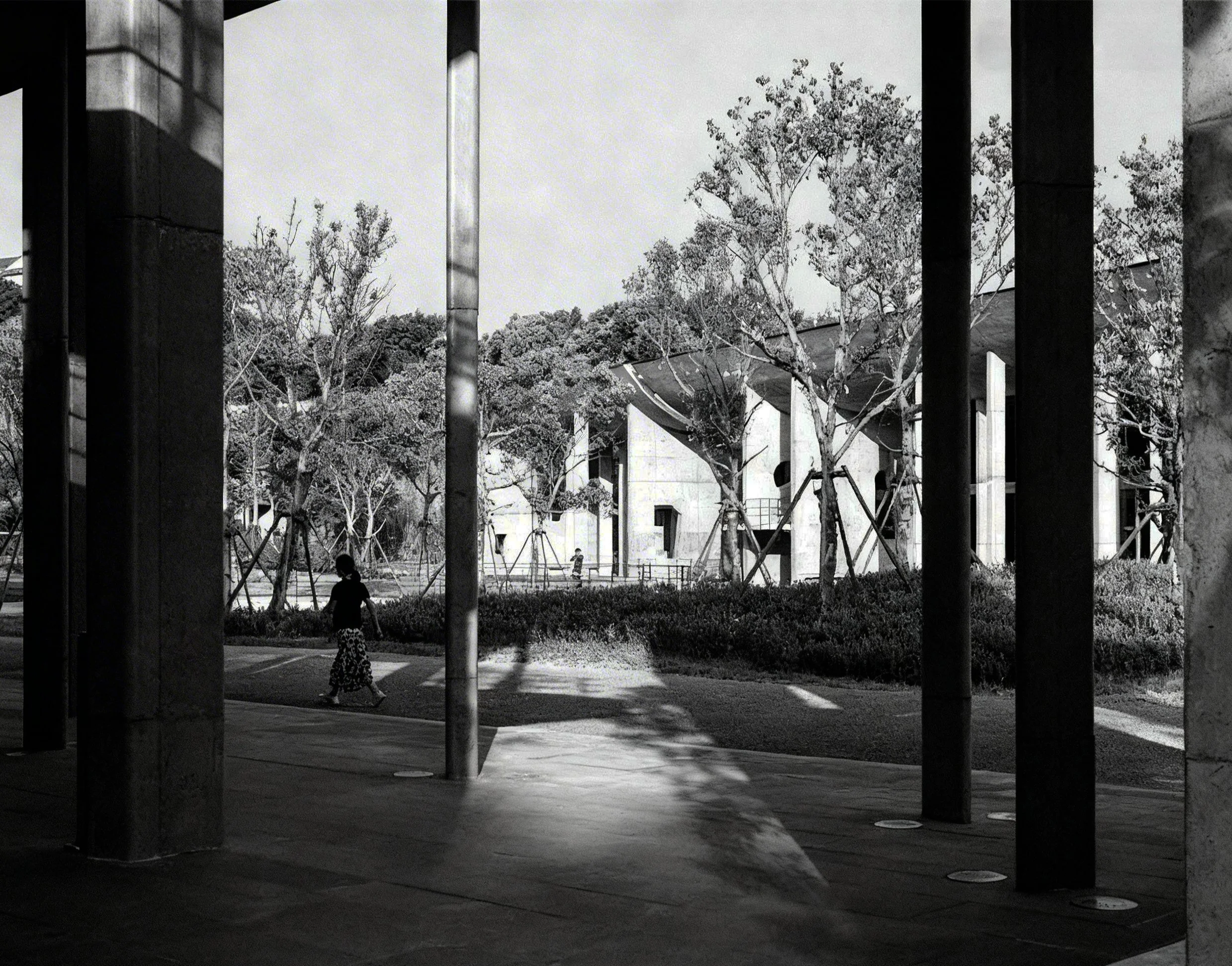 Black and white photo of a modern outdoor area with trees, buildings, and structural columns, casting shadows on the ground with children playing in the background.