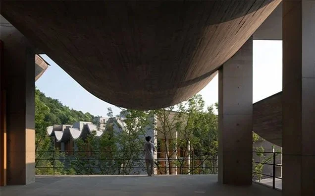 A person stands on a balcony or terrace underneath a large, curved architectural structure with wooden panels, overlooking a scenic view of trees and buildings.