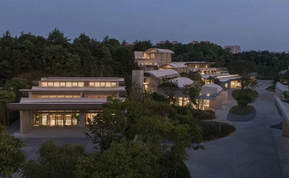 Modern architectural complex with multiple buildings and curved roofs, illuminated at dusk, surrounded by trees and hills.