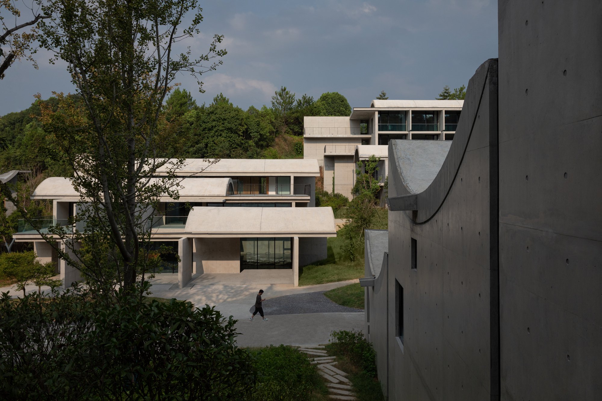 Modern architectural buildings with concrete exteriors, surrounded by greenery and trees, under a partly cloudy sky. A person is walking on a gravel path in the courtyard.