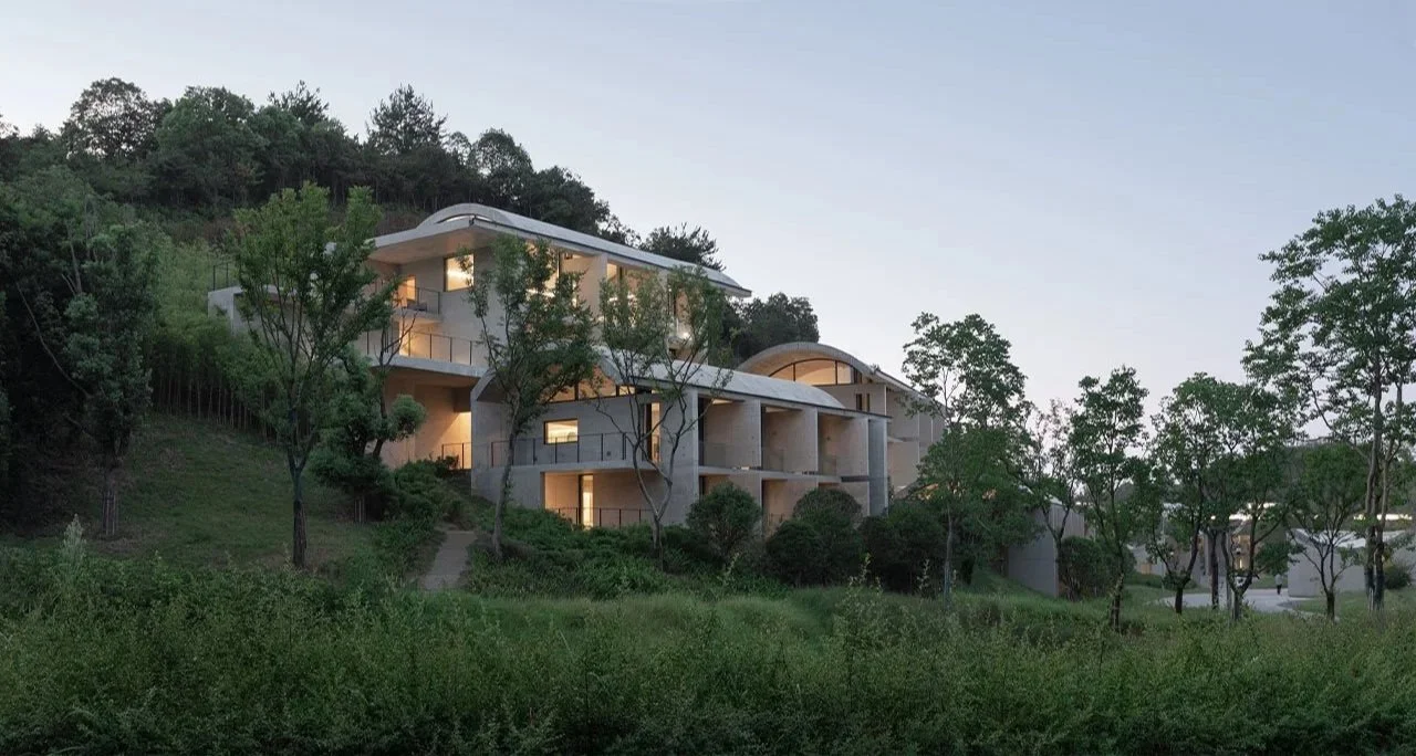 Modern multi-story house with large windows built on a hillside with trees and greenery during dusk.