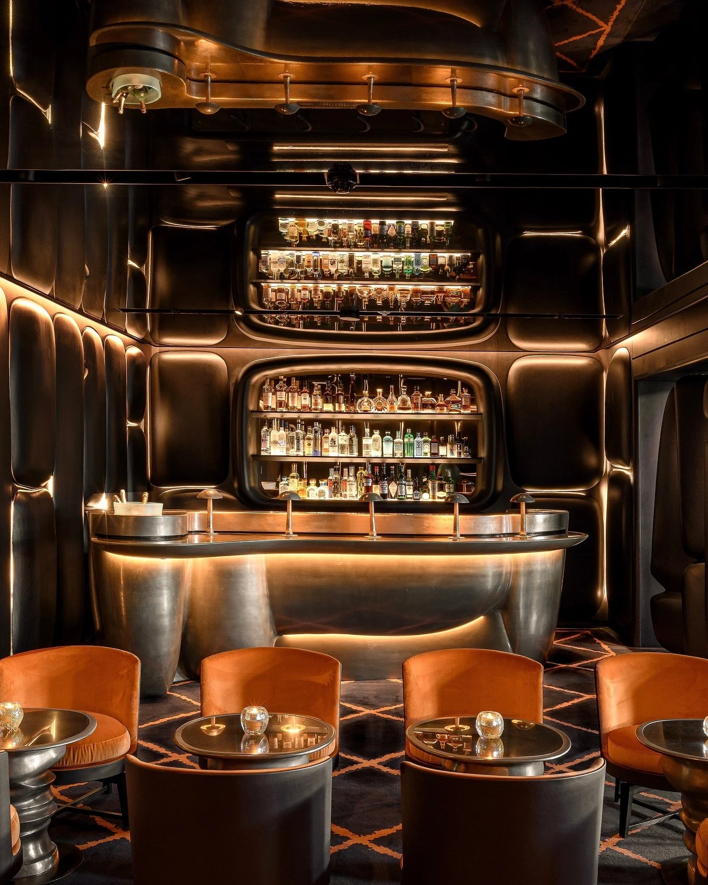 A bar with a sleek, modern design featuring a curved metallic counter, and illuminated shelves stocked with various liquor bottles behind it. The area is dimly lit with warm lighting and has wooden chairs with rounded backs and small tables with candles in glasses in front.