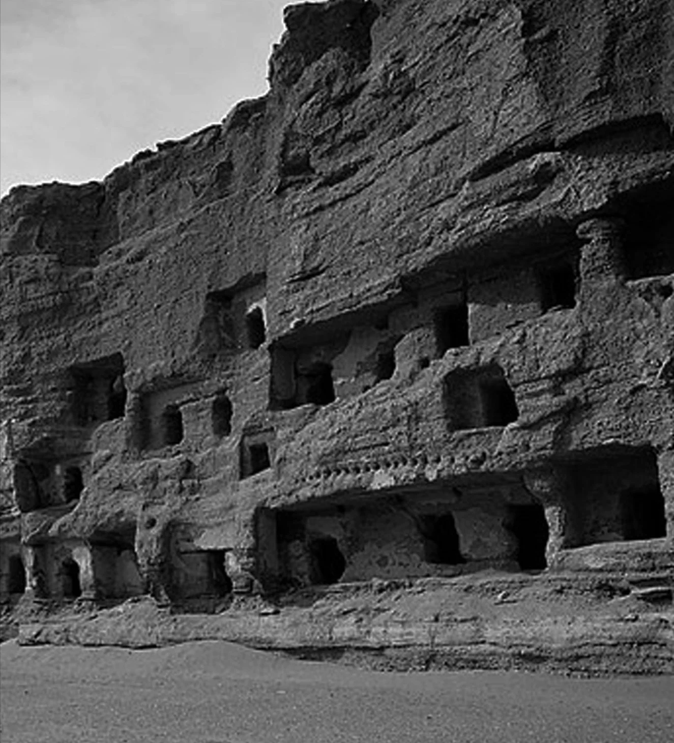 Black and white photo of an ancient rock-cut structure with multiple rectangular openings carved into a cliffside.