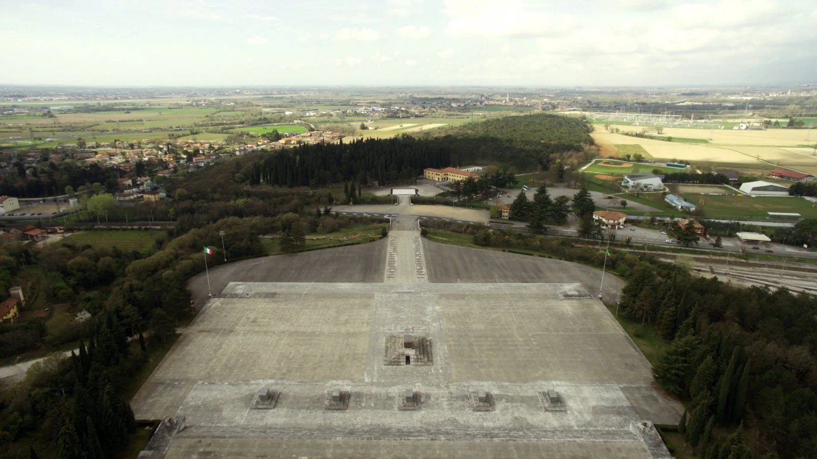 Aerial view of a large memorial with a wide staircase leading to a flat platform, flanked by two flagpoles with the Italian flags, surrounded by trees and fields, with a cityscape and farmland in the background.