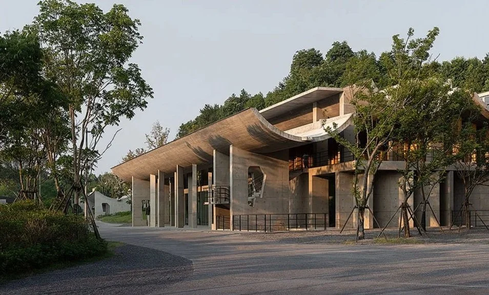 Modern building with curved roof and large windows, surrounded by trees and a paved path.