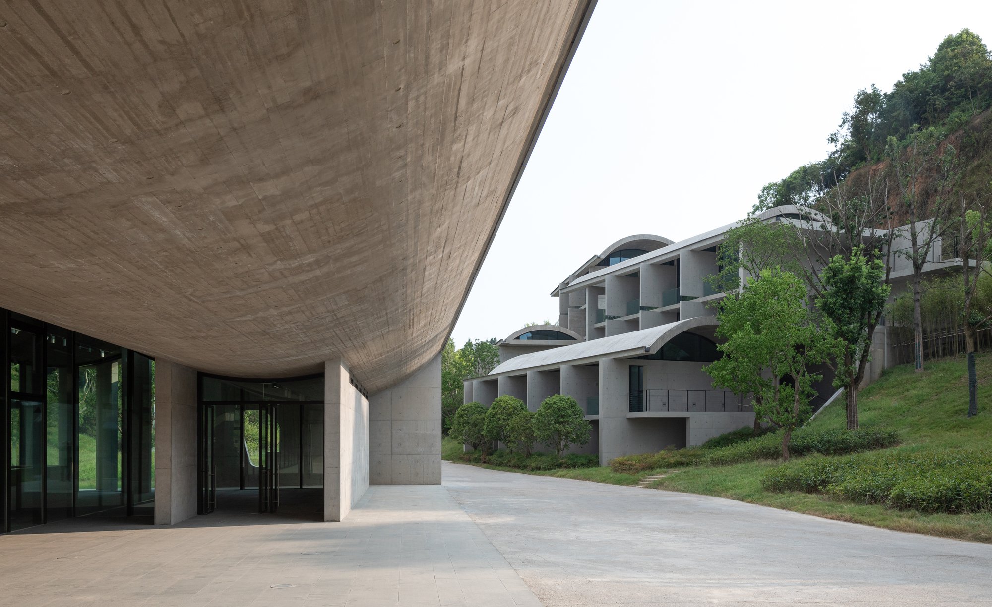 Modern architectural building with a large overhanging concrete roof, glass doors, and multiple residential units on a hillside, surrounded by green trees.