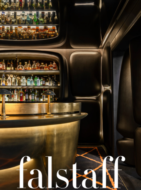 A bar with shelves of liquor bottles behind a round gold-colored counter and modern black seating in a dimly lit, stylish setting.